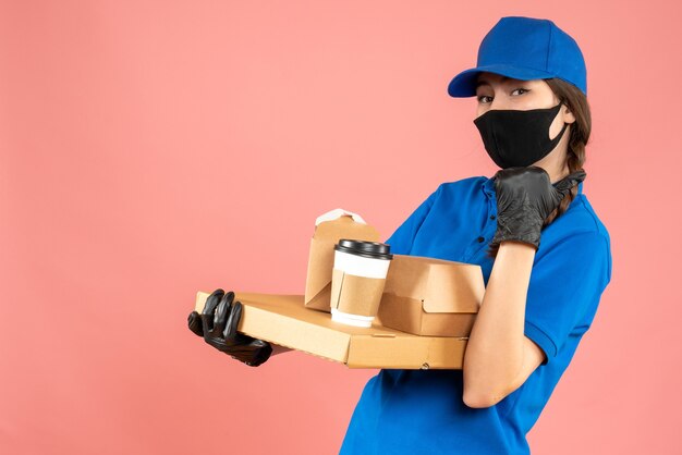 Foto de medio cuerpo de una chica de mensajería con máscara médica y guantes con órdenes apuntando hacia atrás sobre fondo de melocotón pastel