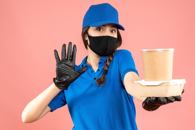 Foto de medio cuerpo de una chica de mensajería con máscara médica y guantes sosteniendo una pequeña caja de café que muestra cinco sobre fondo melocotón pastel