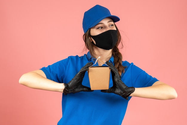 Foto gratuita foto de medio cuerpo de una chica de mensajería ocupada con máscara médica y guantes sosteniendo una pequeña caja posando para la cámara sobre fondo melocotón pastel