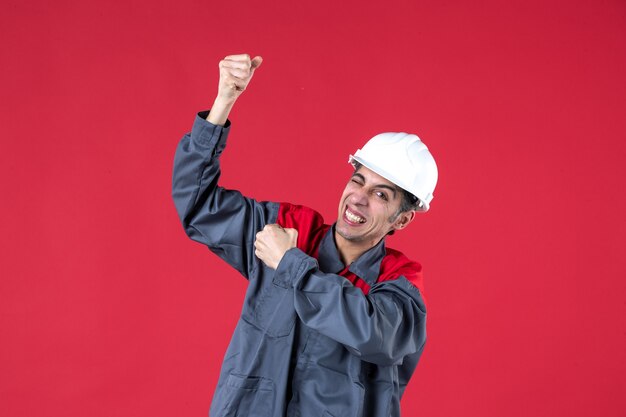Foto de medio cuerpo del joven constructor confiado en uniforme con casco mostrando su músculo en la pared roja aislada