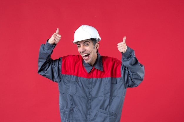 Foto gratuita foto de medio cuerpo de sonriente joven constructor divertido en uniforme con casco apuntando hacia arriba en la pared roja aislada