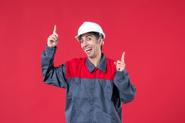 Foto de medio cuerpo de sonriente joven constructor en uniforme con casco apuntando hacia arriba en la pared roja aislada