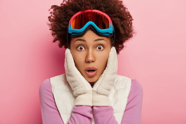 Foto de miradas femeninas rizadas emocionales estupefactas con expresión de cara de sorpresa en la cámara, mantiene ambas manos en las mejillas, usa gafas de esquí para hacer snowboard, aislado sobre la pared rosada del estudio