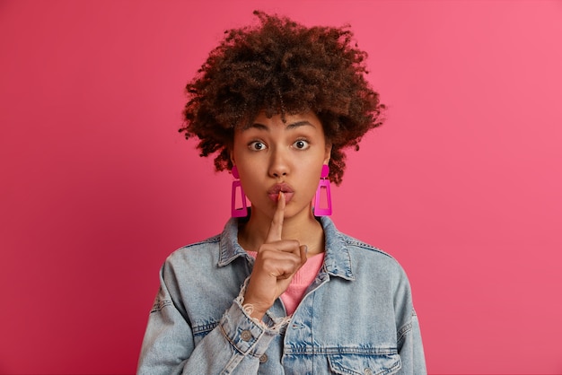 Foto gratuita foto de una misteriosa joven que tiene un plan secreto, hace un gesto de silencio, presiona el dedo índice contra los labios, dice que guarde silencio, prohíbe hablar, usa aretes rosas y una chaqueta de mezclilla, exige silencio