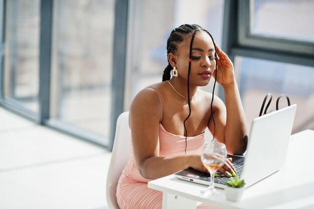 Foto de moda de una joven y hermosa mujer afroamericana romántica con una copa de vino usando un vestido rosa trabajando con una laptop