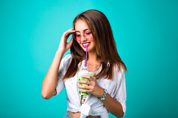 Foto de moda de primavera de estudio de niña sonriente, estilo retro hippie, bebiendo fondo de menta batido sabroso, chica hipster alegre feliz disfruta de su cóctel, estado de ánimo positivo.