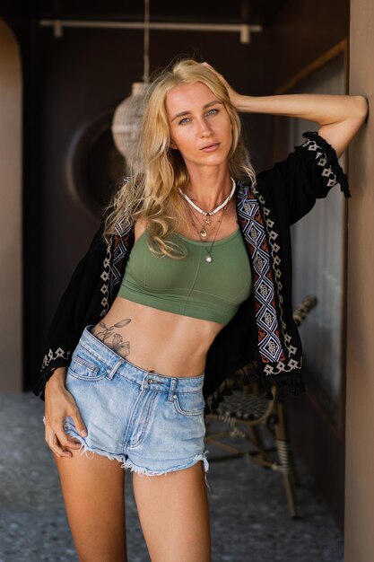 Foto de moda de verano de mujer rubia seductora con figura perfcet en top sexy y jeanse posando al aire libre. Luciendo complementos boho.