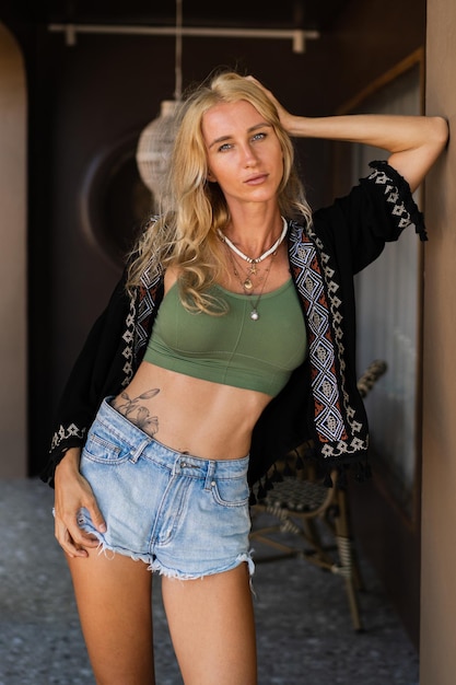 Foto de moda de verano de mujer rubia seductora con figura perfcet en top sexy y jeanse posando al aire libre. Luciendo complementos boho.