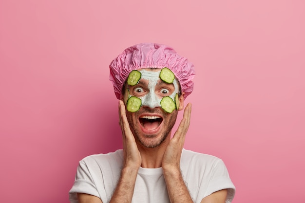 Foto de modelo masculino divertido que toca las mejillas, se ríe alegremente, disfruta de la frescura de la piel, recibe tratamientos de belleza regulares, usa mascarilla facial con rodajas de pepino