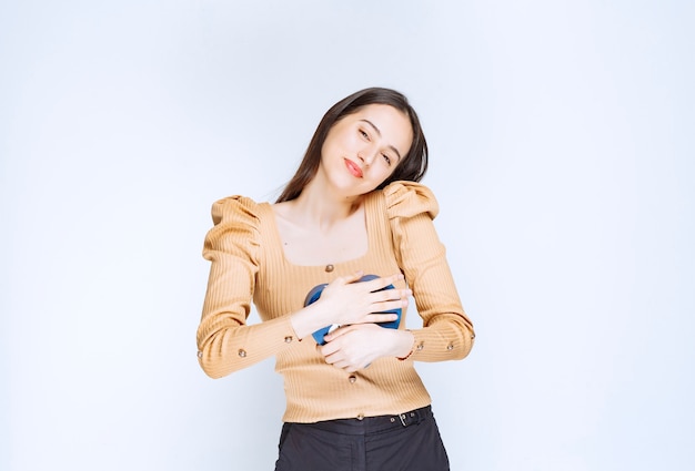 Foto de un modelo de mujer joven abrazando una caja de regalo en forma de corazón contra la pared blanca.