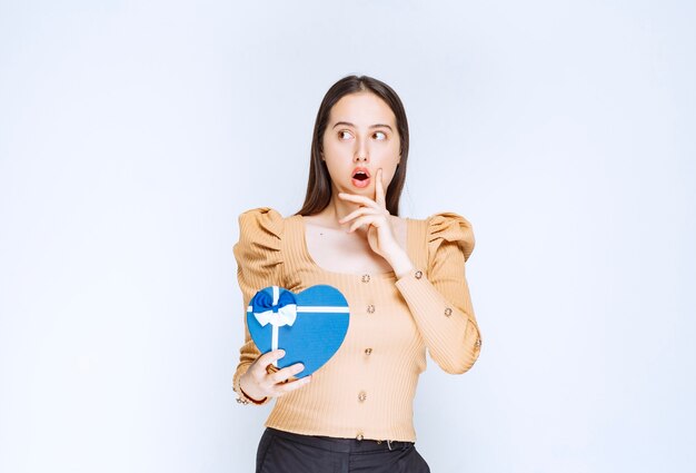 Foto de un modelo de mujer joven sosteniendo una caja de regalo en forma de corazón contra la pared blanca.