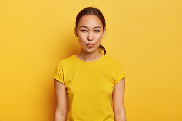 Foto monocromática de mujer hermosa con apariencia asiática, piel sana, piercing en la oreja, mantiene los labios doblados, espera un beso, tiene un estado de ánimo coqueto, viste una camiseta casual amarilla. Concepto de expresiones faciales