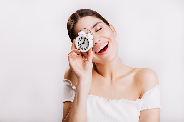 Foto de morena sonriente sin maquillaje posando con reloj en pared blanca.