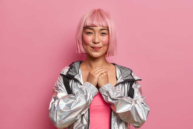 Foto gratuita foto de una mujer adolescente tímida sonriente y complacida que tiene una peluca de cabello rosa con flecos, mantiene las manos juntas, anticipa que suceda algo bueno, vestida con una chaqueta plateada de moda, modelos de interior