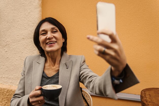 Una foto de una mujer adulta de piel clara sonriente que usa su teléfono móvil mientras toma una taza de café fuera del concepto de tecnología Gadget