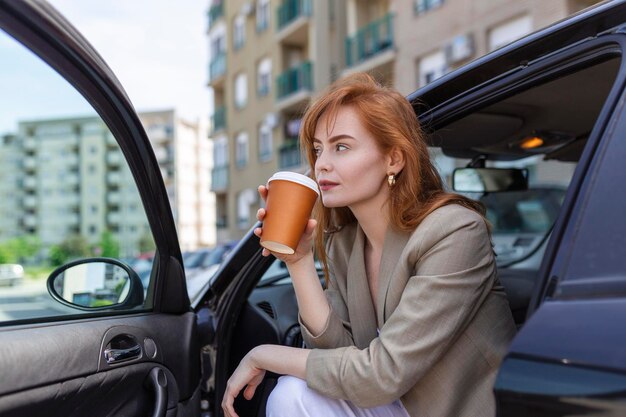 Una foto de una mujer adulta sentada en su auto con la puerta abierta contemplando y bebiendo café de una taza de viaje