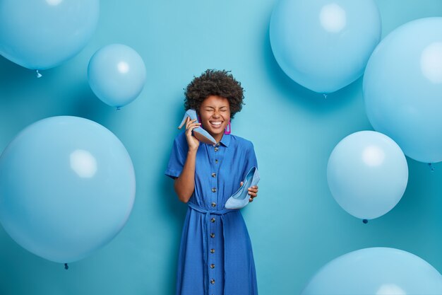 Foto de una mujer afroamericana alegre y exagerada que se ríe y se divierte antes de vestirse en una fiesta disco, imita una conversación telefónica, sostiene zapatos de tacón alto cerca de las orejas, aislado en una pared azul