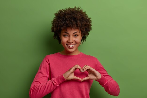 Foto de una mujer afroamericana bonita que expresa amor, está en un estado de ánimo romántico, muestra el signo del corazón, confiesa sentimientos sinceros, tiene simpatía, se viste con un jersey rosado, posa contra la pared verde