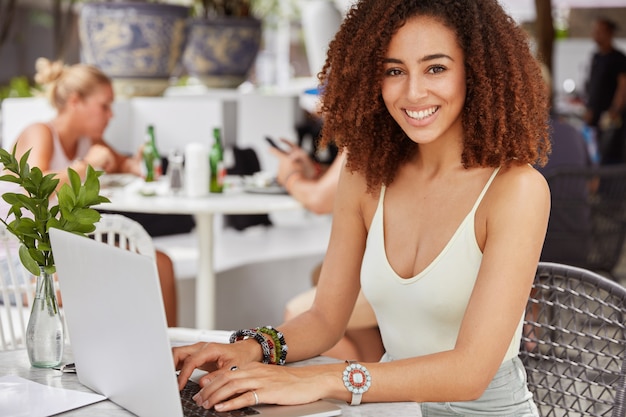 Foto de mujer afroamericana complacida con amplia sonrisa brillante, vestida de manera informal, teclados en la computadora portátil