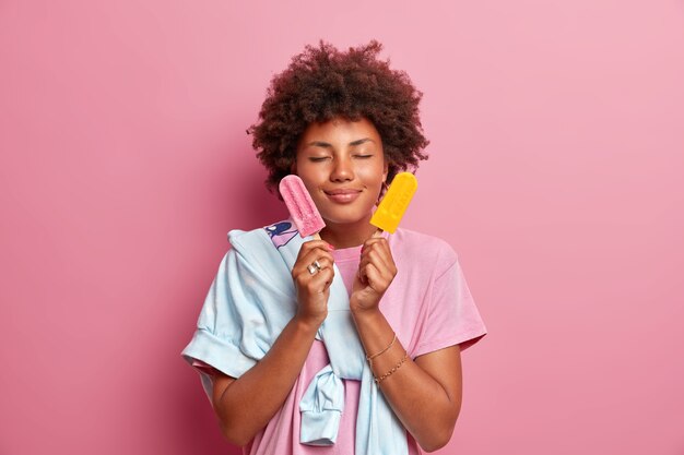 Foto de mujer afroamericana complacida se encuentra con los ojos cerrados sostiene dos helados