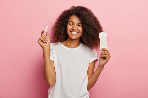 Foto de mujer afroamericana encantada sostiene tampón y toalla sanitaria, vestida con camiseta blanca, aislada sobre pared rosa. Mujeres, pms
