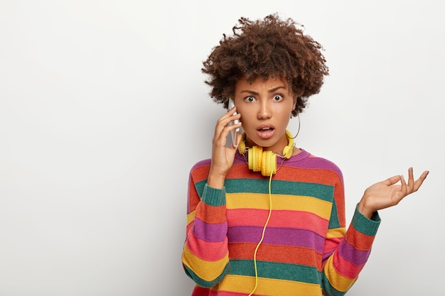 Foto de mujer afroamericana indignada y perpleja levanta la palma, habla por celular, vestida con ropa casual, parece infeliz, posa sobre un fondo blanco.