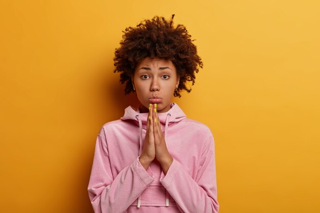 Foto de una mujer afroamericana insatisfecha que mantiene las palmas juntas, reza o suplica, ruega por tu ayuda, frunce los labios, mira con tristeza, posa contra la pared amarilla, viste una sudadera con capucha rosada informal