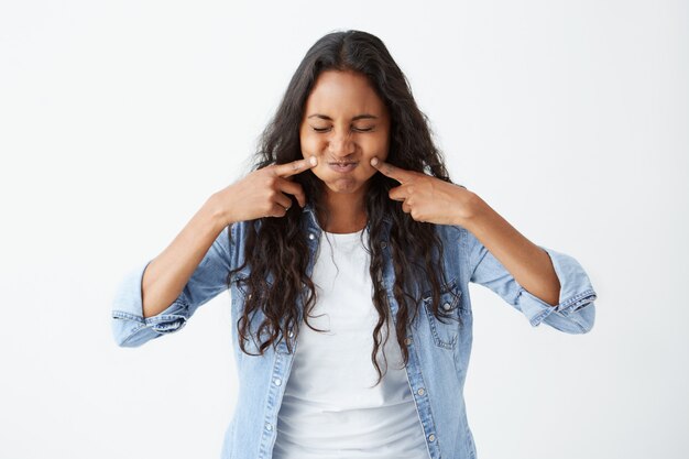 foto de una mujer afroamericana morena vestida casualmente con los ojos cerrados haciendo una mueca que sopla sus mejillas tocándolas con los dedos, con expresión complacida.