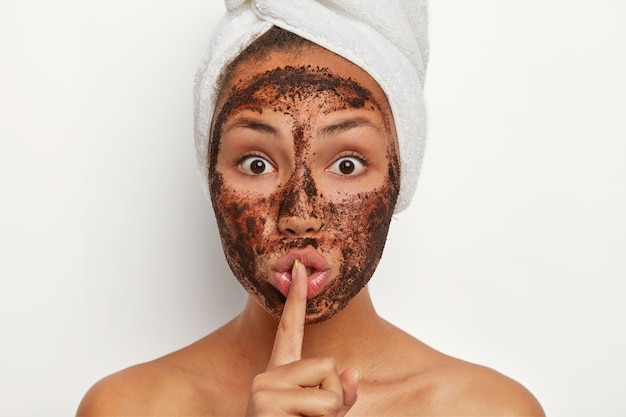 Foto de mujer afroamericana sorprendida con exfoliante de café para pelar los poros, hace un gesto de silencio, tiene expresión facial de sorpresa, muestra los hombros desnudos, dice un secreto, tiene un procedimiento de cosmetología