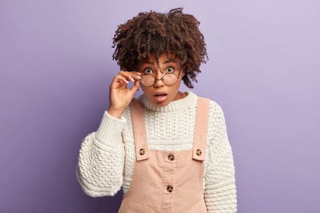 La foto de una mujer afroamericana sorprendida mantiene la mano en el marco de las gafas, mira fijamente con sorpresa directamente, usa un jersey blanco con un mono, expresa asombro mientras escucha noticias impresionantes