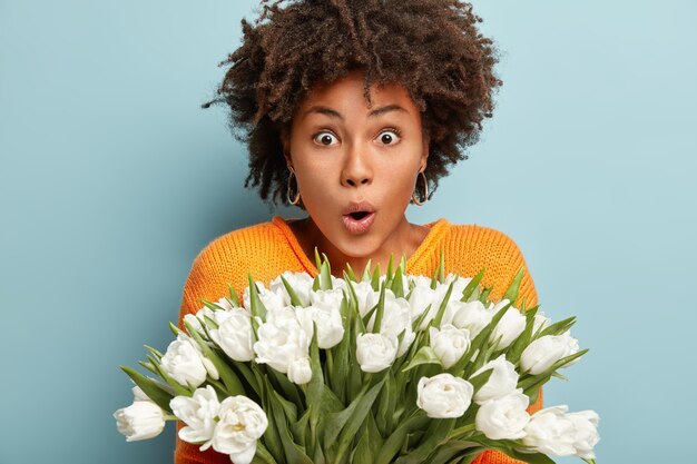 Foto de mujer afroamericana sorprendida con la respiración contenida, no puedo creer los ojos que recibió un ramo tan grande de flores primaverales, abre la boca del shock, aislado sobre una pared azul. Wow, que tulipanes