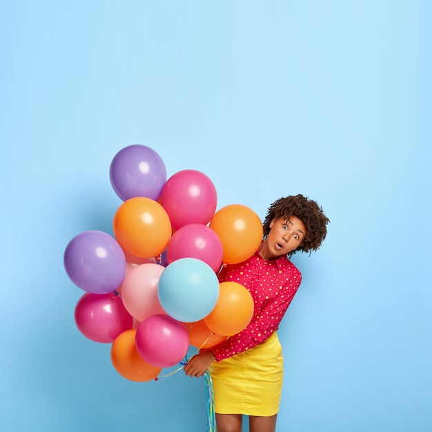 La foto de una mujer afroamericana sorprendida se ve con expresión omg, sostiene muchos globos de aire de helio coloridos, vestida con una camisa y una falda vívidas