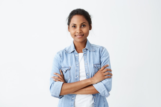 La foto de la mujer afroamericana tranquila con el pelo negro que se coloca cruzó las manos que tenían una sonrisa sincera y encantadora que presentaba en la pared blanca. Feliz mujer de piel oscura regocijándose de su vida.