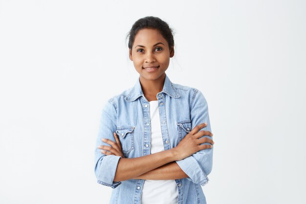 La foto de la mujer afroamericana tranquila con el pelo negro que se coloca cruzó las manos que tenían una sonrisa sincera y encantadora que presentaba en la pared blanca. Feliz mujer de piel oscura regocijándose de su vida.