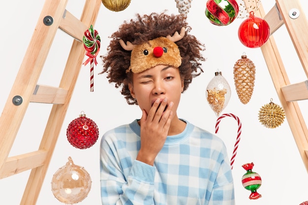 Foto de mujer agotada que se despierta muy temprano en la mañana bosteza y quiere dormir tiene mucho que ver antes de Año Nuevo vestida con pijama a cuadros y antifaz para dormir decora la casa con adornos navideños