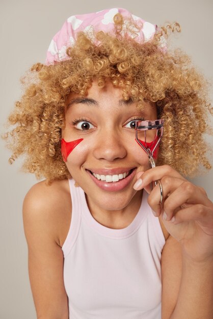 La foto de una mujer alegre de cabello rizado usa un rizador de pestañas usa un gorro de baño y una camiseta casual aplica parches de belleza rojos debajo de los ojos se somete a un tratamiento de cuidado de la piel se prepara para la cita aislada sobre una pared gris