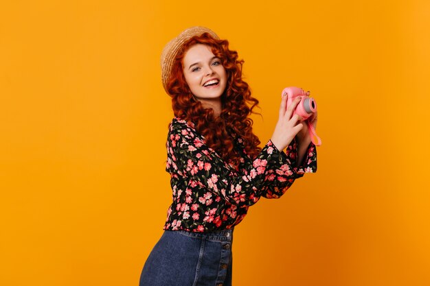 Foto de mujer alegre con ojos azules, tomando fotos con cámara rosa. Chica con sombrero de paja, blusa y falda sonriendo.