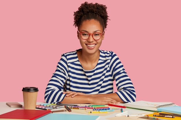 Foto gratuita foto de mujer alegre de piel oscura con cabello afro peinado en cola de caballo, tiene una sonrisa con dientes, está contenta con un buen resultado del trabajo, crea una imagen en el bloc de notas, usa anteojos con montura roja, aislado en rosa