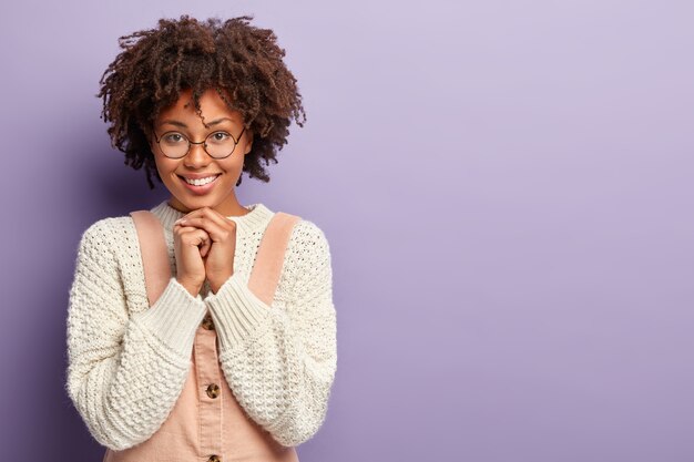 Foto de mujer alegre con piel oscura, muestra sus dientes, mantiene las manos debajo de la barbilla, usa un jersey blanco de gran tamaño, posa sobre una pared violeta con un espacio en blanco para su contenido publicitario. Concepto de emociones