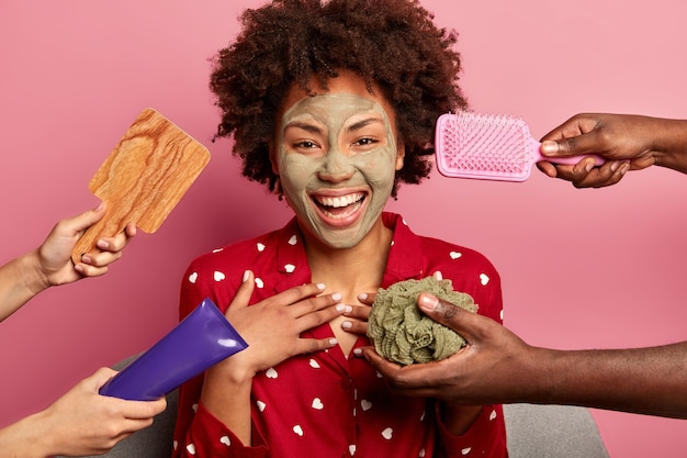 Foto de mujer alegre de piel oscura que disfruta de tratamientos de belleza, se ha aplicado mascarilla de arcilla en la cara, usa pijama, rodeada de peine