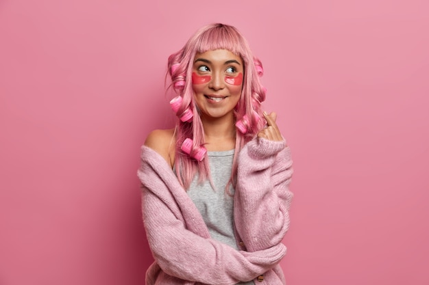 Foto de mujer alegre y soñadora hace mini gesto de corazón símbolo coreano usa parches de colágeno debajo de los ojos, rizadores de pelo