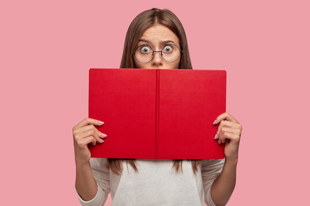 Foto de mujer ansiosa de cabello oscuro que oculta la cara con el libro, usa anteojos redondos, suéter blanco, lee información sorprendente, mira con ojos llenos de miedo, aislada sobre una pared rosa, se siente desconcertado