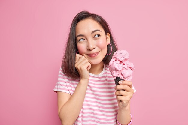 Foto de mujer asiática bonita que se ve soñadora a un lado recuerda agradables recuerdos come sabroso postre de verano sostiene un gran cono de helado vestida con modelos casuales de camiseta