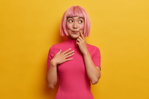 Foto de mujer asiática feliz sonriente con peluca de pelo rosa, imagina algo agradable, se ve con expresión pacífica de ensueño