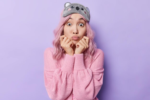 La foto de una mujer asiática sorprendida con piel sana mantiene las manos debajo de la barbilla tiene los labios doblados y los ojos muy abiertos reaccionan ante algo impactante que usa máscara para dormir y un puente rosa aislado sobre un fondo morado