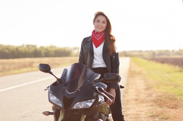 Foto de una mujer de aspecto agradable que el motorista se sienta en una moto negra rápida, lleva un elegante pañuelo rojo y una chaqueta de cuero, viaja solo