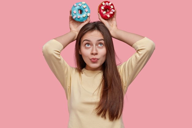 Foto gratuita foto de mujer atractiva con cabello oscuro, mantiene coloridos donuts sobre la cabeza, demuestra comida chatarra