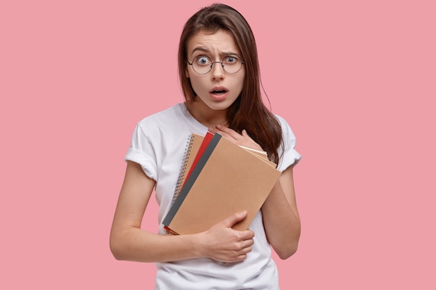Foto de mujer atractiva sorprendida tiene expresión de sorpresa, lleva bloc de notas en espiral, libros de texto, vestida con una camiseta blanca informal