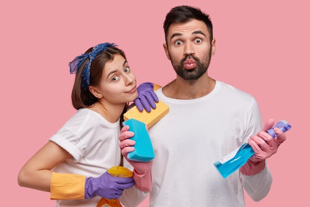 La foto de una mujer atractiva usa diadema, guantes protectores, se para cerca del esposo que dobla los labios, lleva esponjas y detergente, lleva la casa al orden
