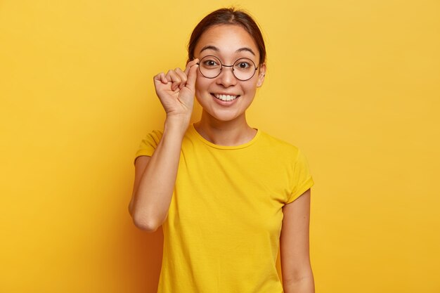 Foto de mujer atractiva se ve curiosamente, tiene expresión feliz, toca el marco de los anteojos, usa camiseta amarilla, lee buenas noticias, concentrada, posa en interiores. Expresiones de rostro humano